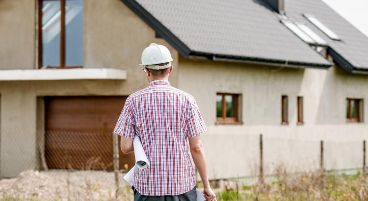 A man in a hard hat holding rolled plans and looking at a house under construction