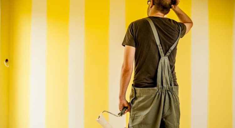 A painter in overalls scratching his head while looking at a yellow and white striped wall